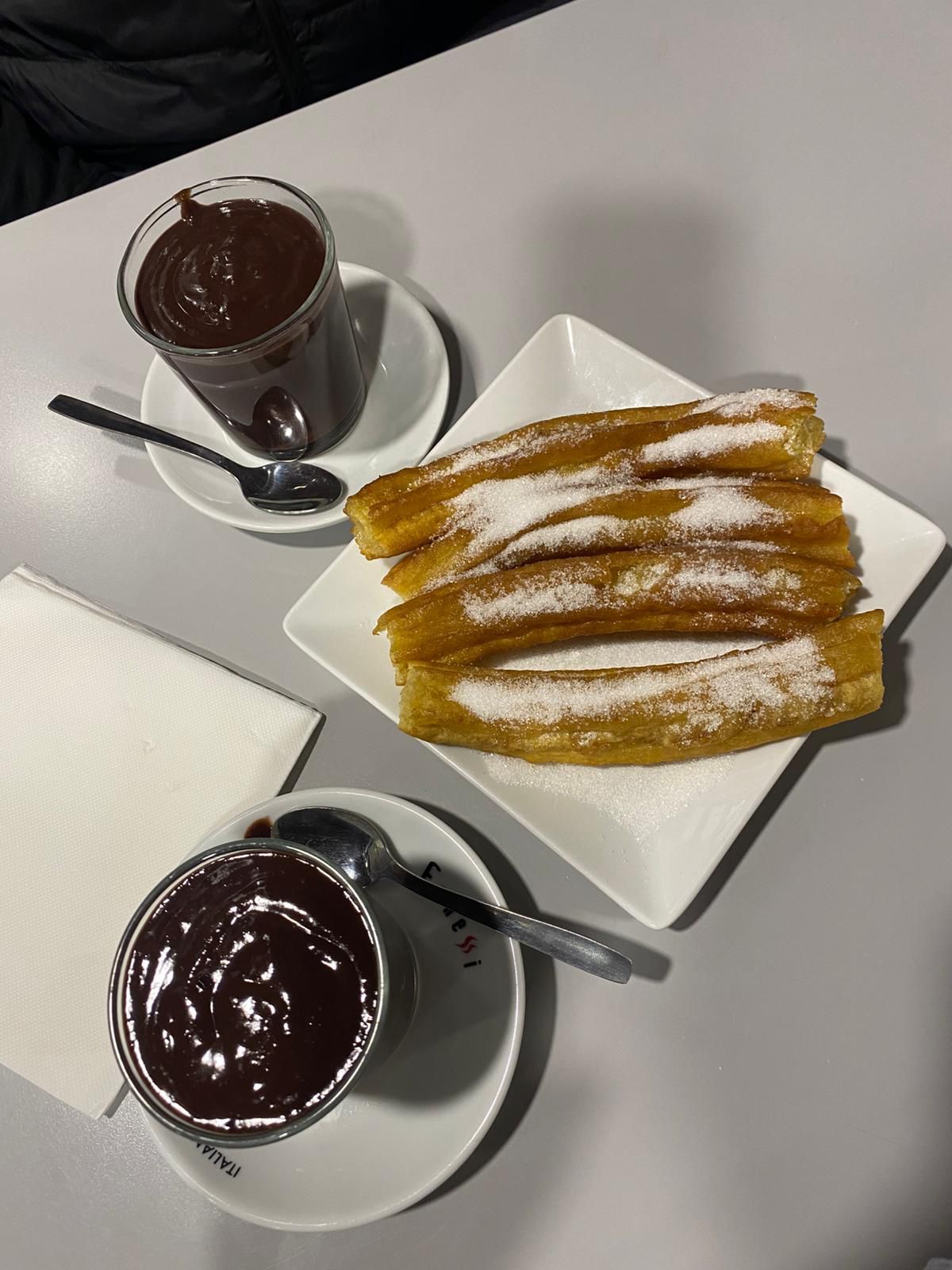 Churros espolvoreados con azúcar sobre un plato blanco, servidos con dos tazas de chocolate caliente en una mesa.