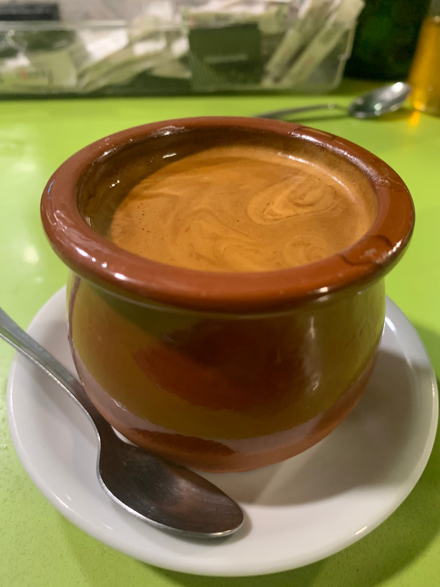 Taza de café en una pequeña cafetera de cerámica marrón sobre un platillo blanco, con una cuchara sobre una superficie verde.