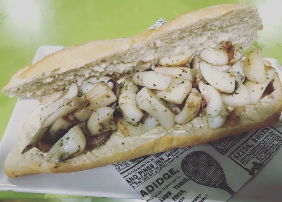 Sándwich de calamares a la plancha en baguette, sobre un plato, con un periódico antiguo debajo.