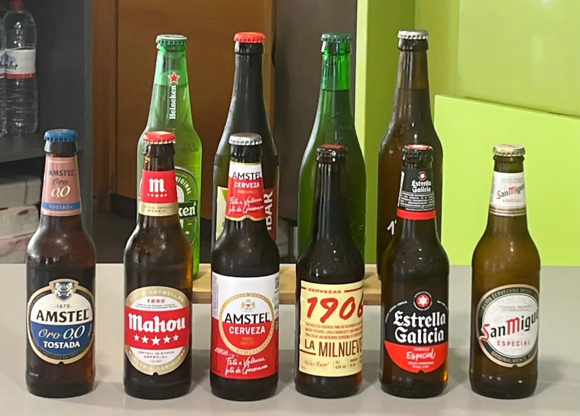 Varias botellas de cerveza alineadas sobre una superficie, cada una con una etiqueta y un color diferente, sobre un fondo verde claro.