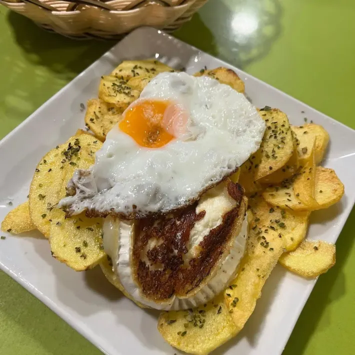 Huevo frito sobre queso Brie frito, servido sobre patatas fritas en un plato blanco.