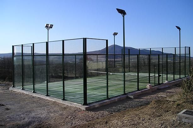 Pista deportiva exterior vallada con superficie verde y luces altas en un paisaje seco y montañoso.