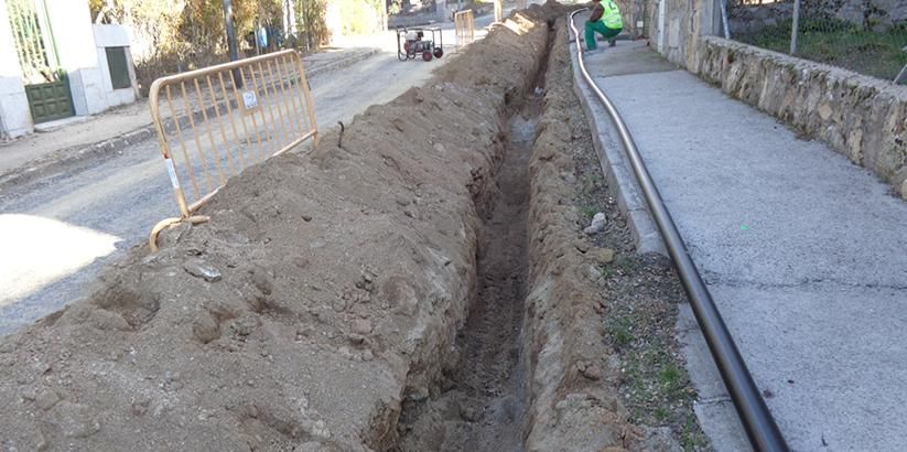 Zanja en construcción junto a una carretera, con tierra amontonada a lo largo del bordillo y trabajadores a lo lejos.