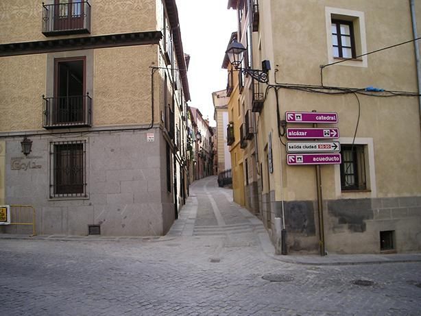 Calle estrecha empedrada entre edificios de color beige con letreros, que asciende hacia la distancia.