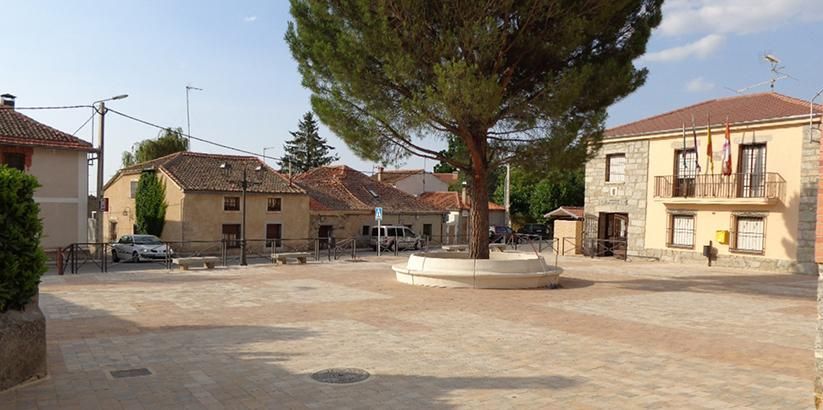 Plaza tranquila del pueblo con un gran árbol en el centro, edificios de piedra y un coche aparcado bajo un cielo despejado.