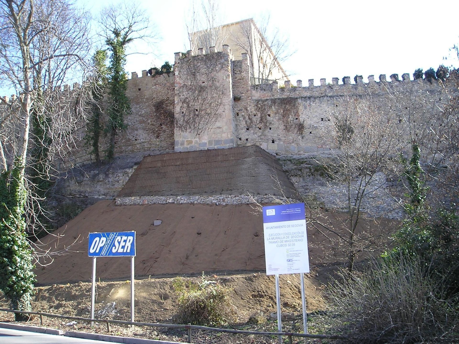 Yacimiento arqueológico situado bajo una muralla de piedra, con señales y una fosa de excavación de tierra.