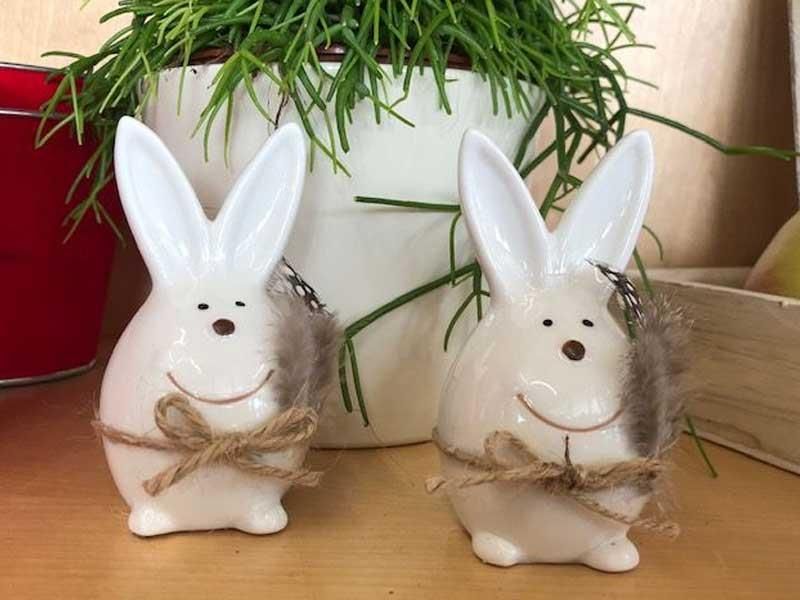 Zwei weiße Hasenfiguren aus Keramik sitzen auf einem Holztisch neben einer Topfpflanze.