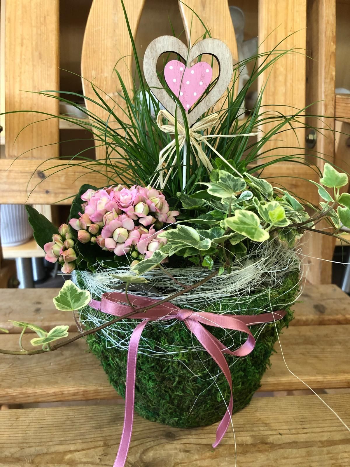 Auf einem Holztisch steht eine Topfpflanze mit Blüten und einem Herz darauf.