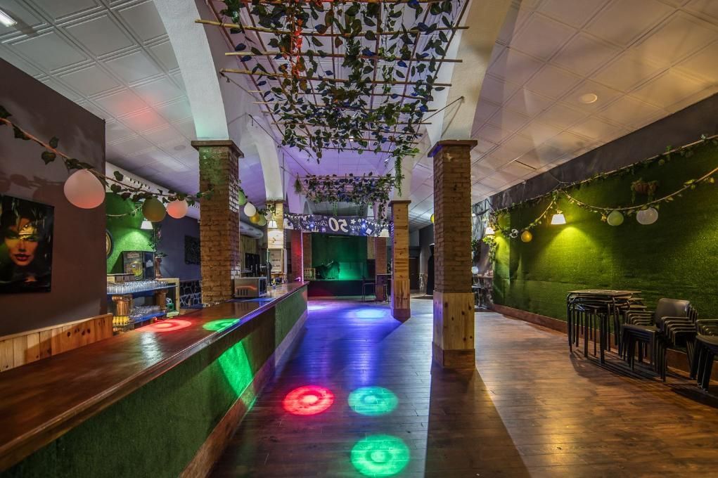 Dentro de un bar/club con pisos de madera, luces verdes y moradas y vegetación decorativa.