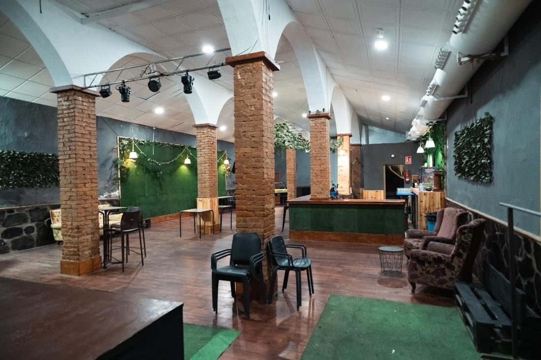 Interior de un local con columnas de ladrillo, techo abovedado, paredes verdes y bar. Se ven zonas de asientos vacías.