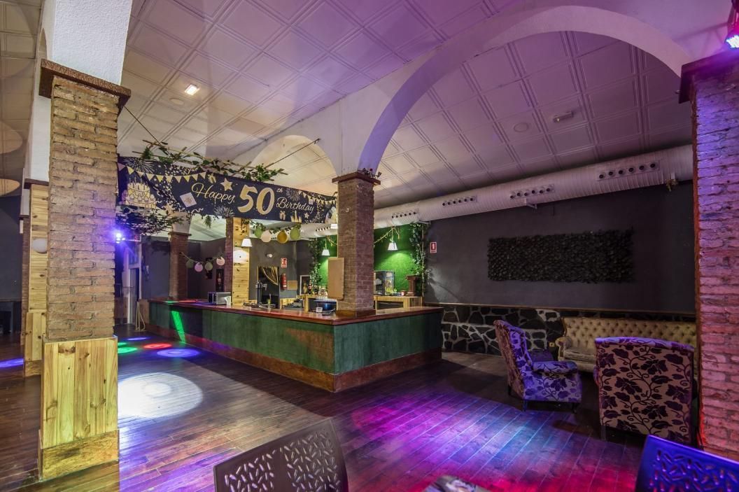 Dentro de un local decorado, bar con arreglo floral, luces de colores en el suelo, paredes en tonos neutros y columnas.