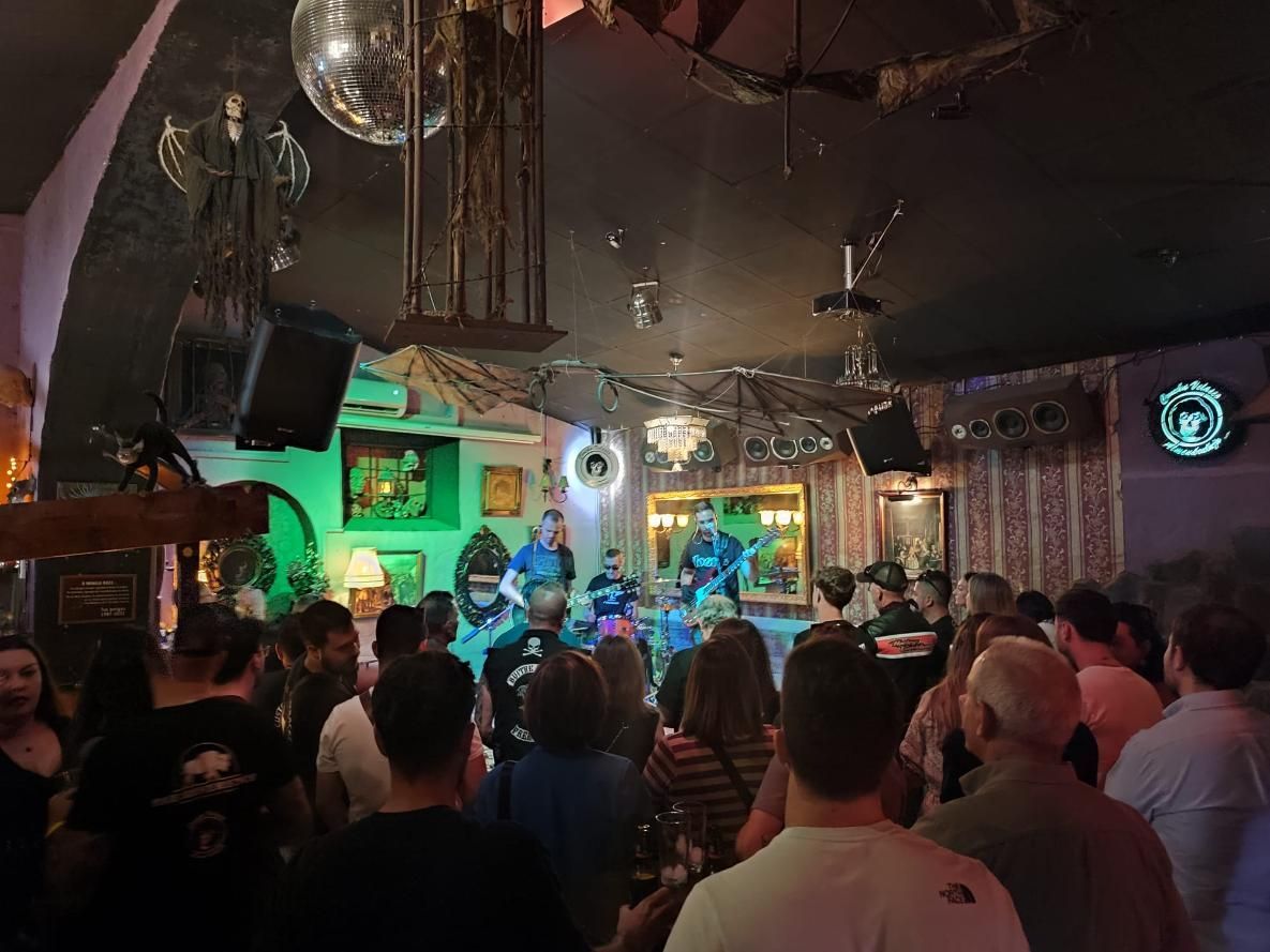 Una banda toca en el escenario en un bar lleno de gente y poco iluminado, con una bola de discoteca y detalles verdes.