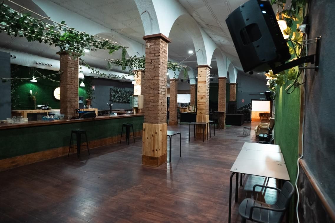 Un espacio vacío para eventos con suelo de madera oscura, columnas, barra y mesas blancas. Vides verdes adornan las columnas.