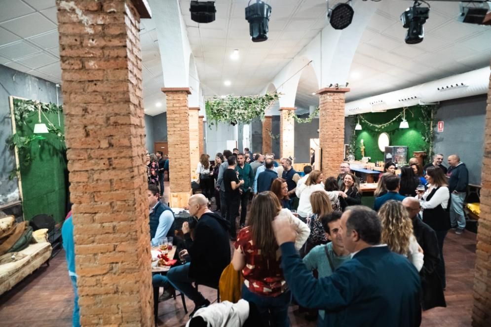 La gente se reúne en un animado evento en interiores. Columnas de ladrillo, plantas y luces decoran el espacio.