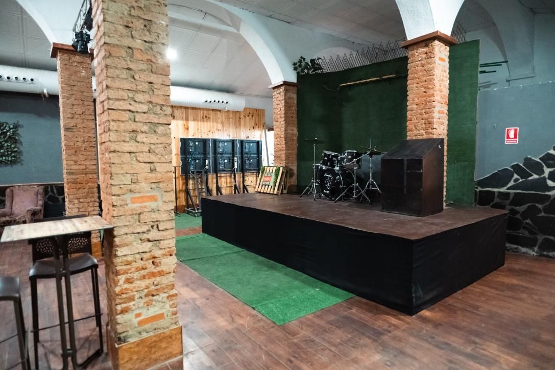 Vista interior de un local de música con escenario, columnas de ladrillo y zona de bar.