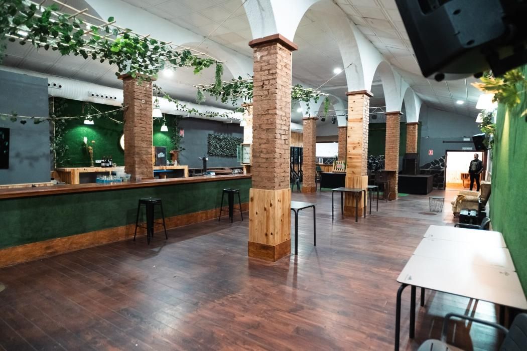 Local interior con suelo de madera, columnas, barra y mesas. Tonos verdes y marrones.