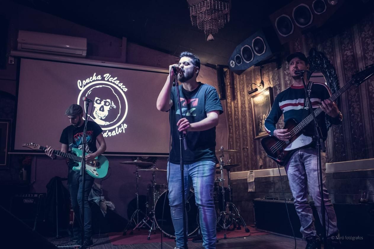 Banda tocando en un bar con poca luz. Cantante, guitarrista y baterista frente a una pantalla con un logo.