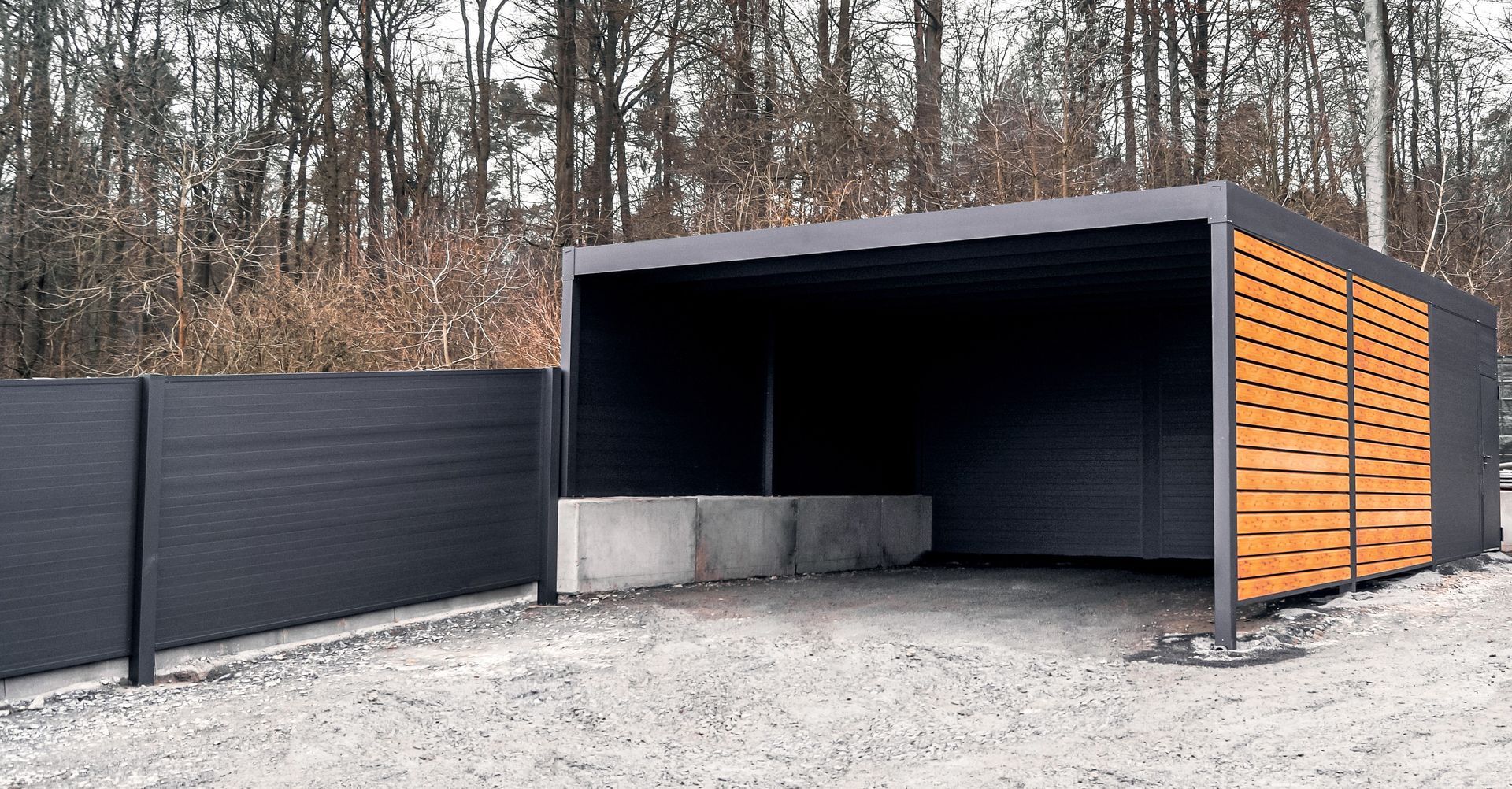 Moderner schwarzer Carport mit seitlichen Holzlamellen auf einer geschotterten Auffahrt in der Nähe eines Waldgebiets