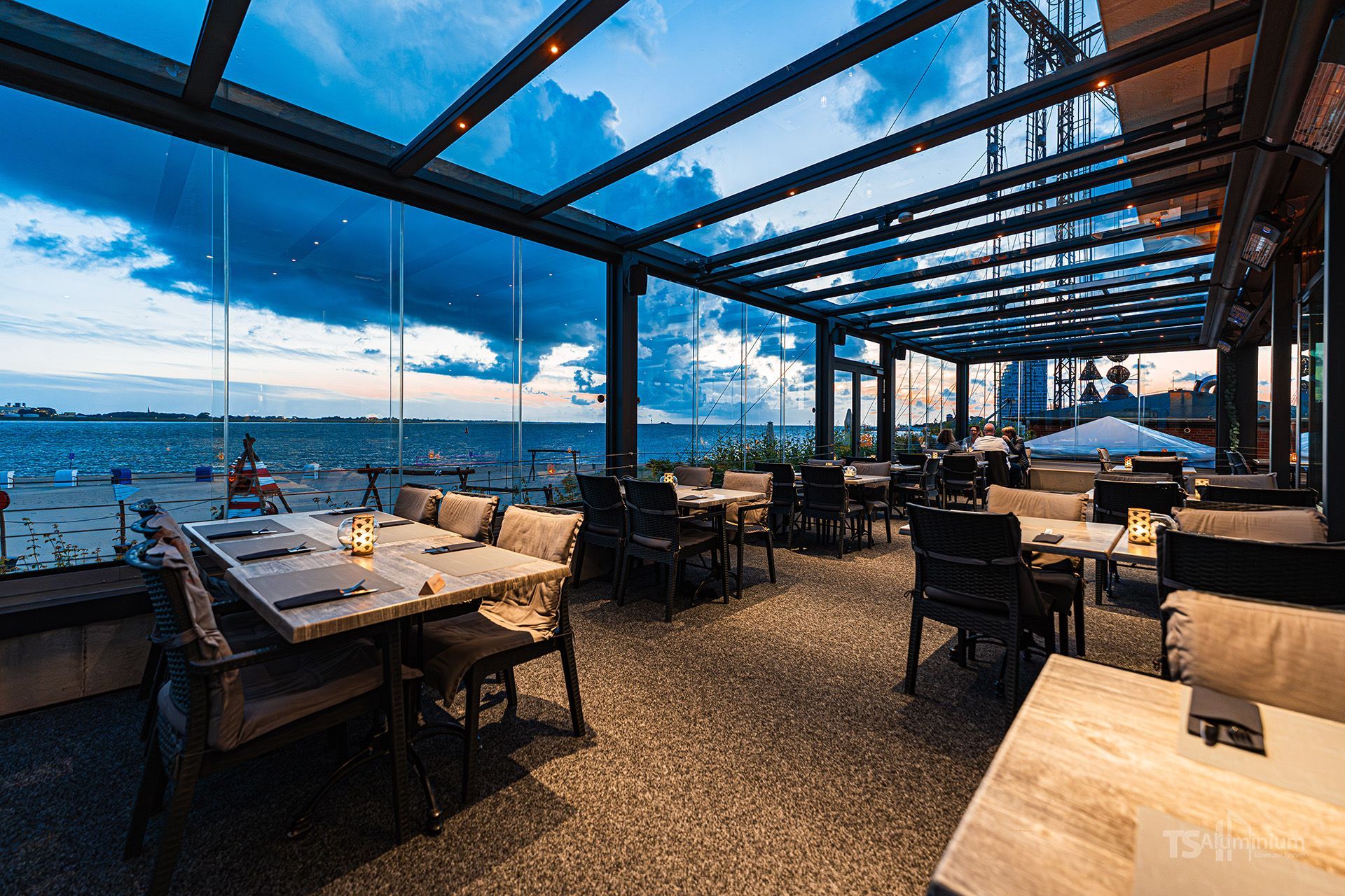 Restaurant am Wasser mit Tischen unter einem Glasdach und Blick aufs Meer bei Sonnenuntergang