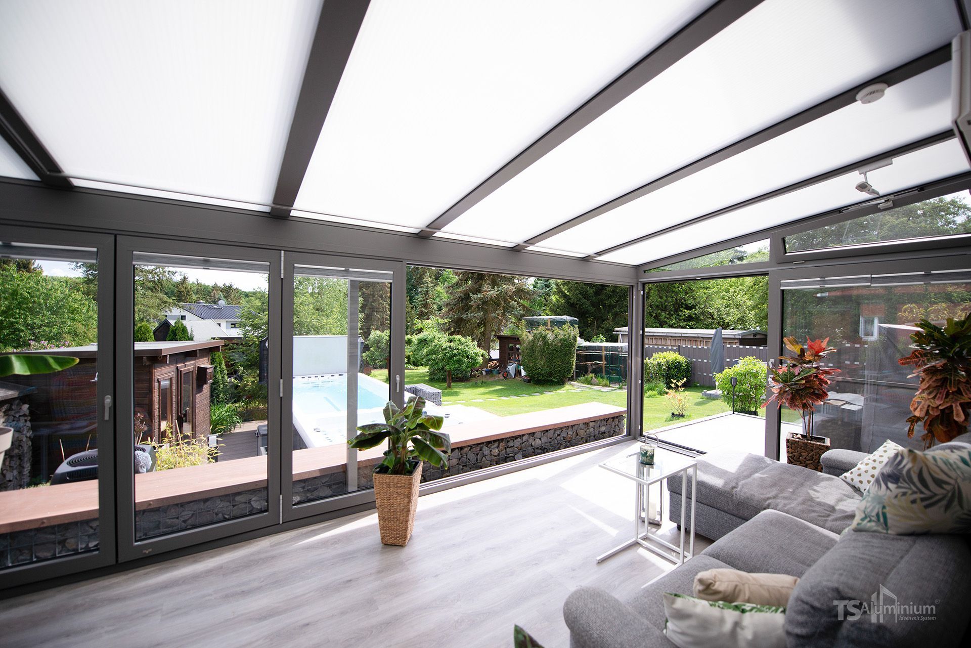 Heller, moderner Wintergarten mit Glastüren, Dachfenster, grauem Sofa und Blick auf den Garten.