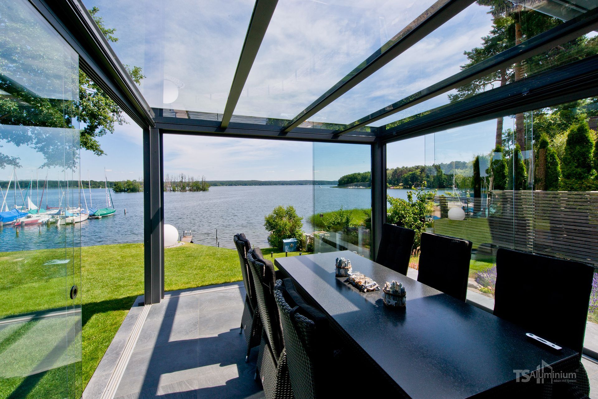 Überdachter Esstisch auf der Terrasse mit Blick auf einen See, einen Rasen und Bäume an einem sonnigen Tag