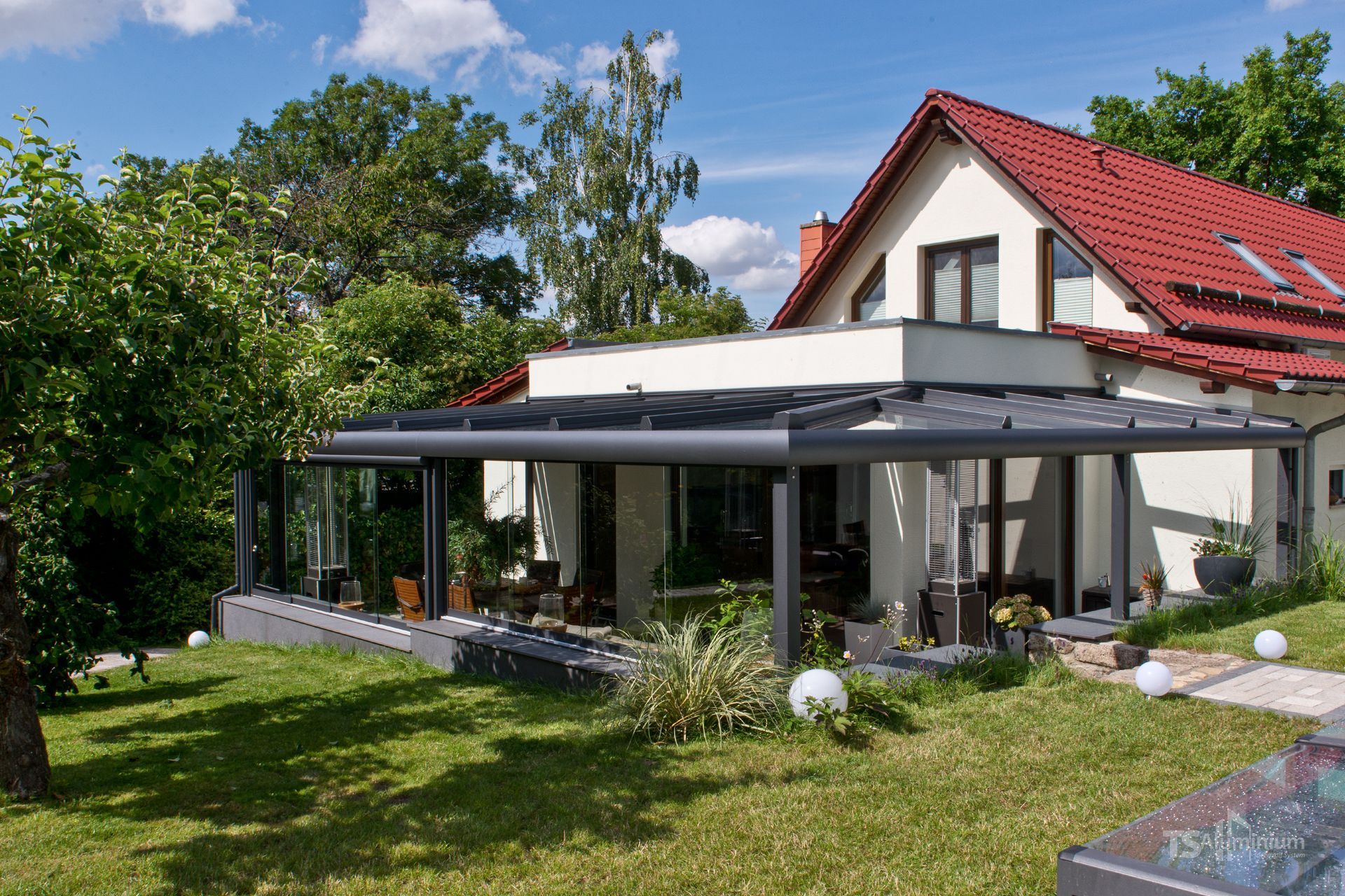 Modernes Haus mit rotem Dach, verglastem Wintergarten und üppig grünem Garten an einem sonnigen Tag