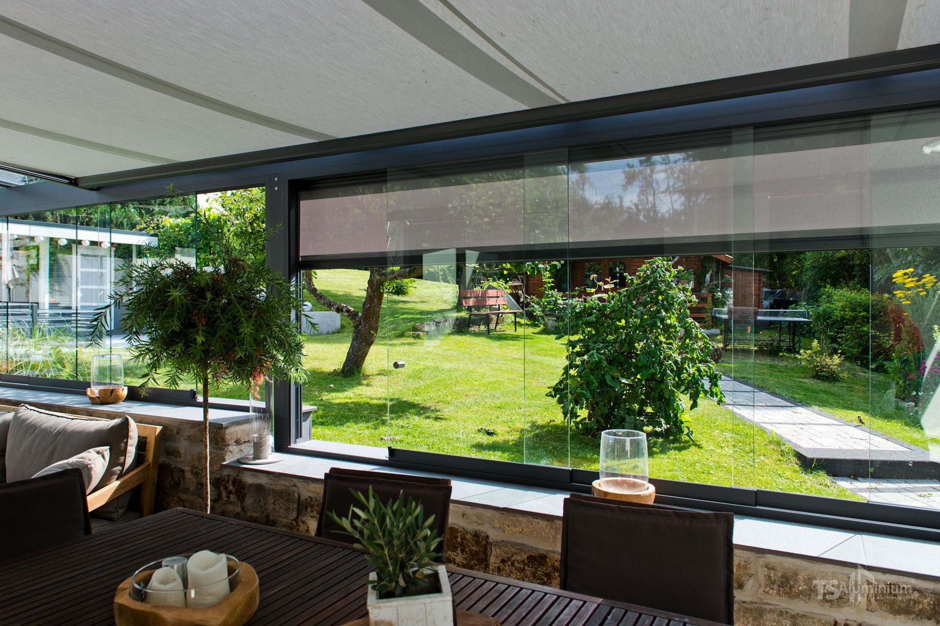 Überdachter Essbereich auf der Terrasse mit Blick auf einen sonnigen Garten und eine Rasenfläche, im Hintergrund Bäume und ein Wohngebäude