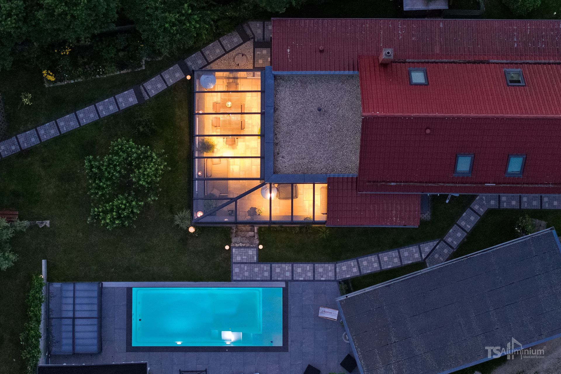 Luftaufnahme eines modernen Hauses in der Abenddämmerung mit beleuchtetem Glasweg, rotem Dach und blauem Swimmingpool.