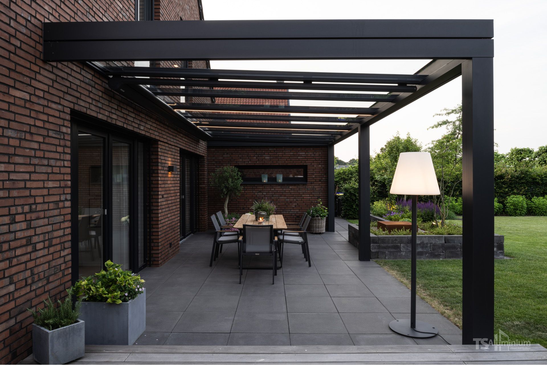 Überdachte Terrasse mit Esstisch im Freien, Pergolabalken, Backsteinwand des Hauses und einer Stehlampe.