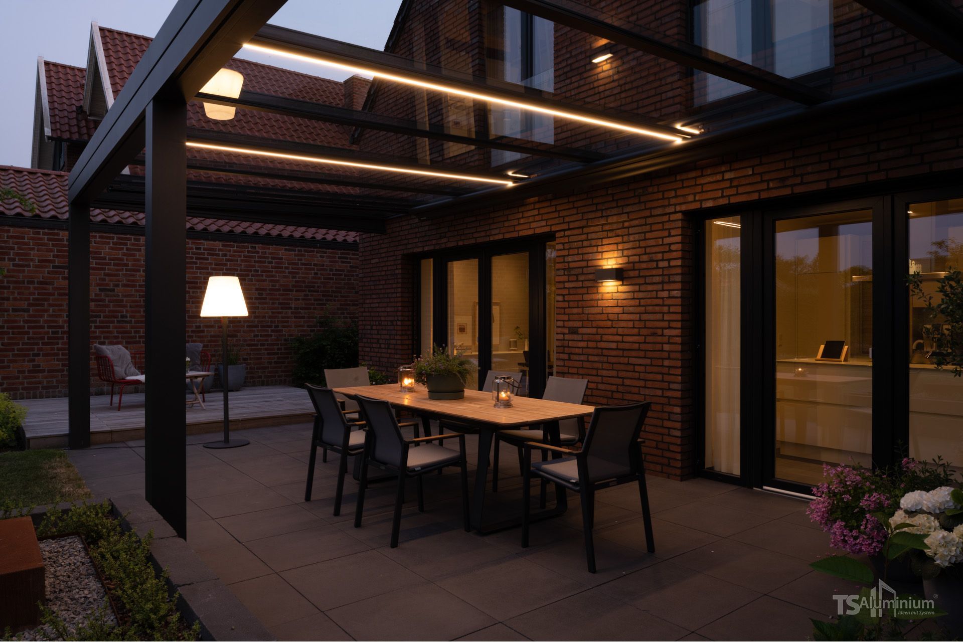 Überdachte Terrasse mit Esstisch, Stühlen und warmem Licht neben einem Backsteinhaus in der Abenddämmerung