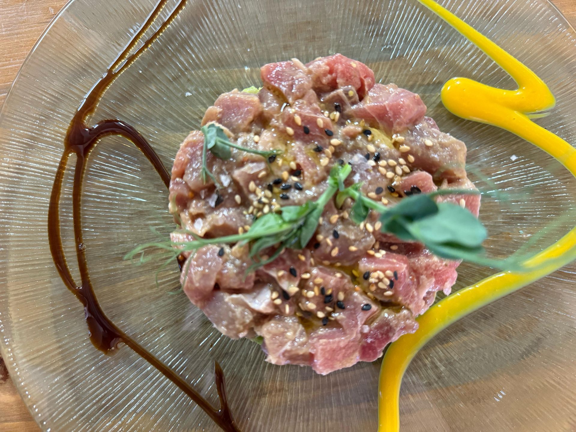 Tartar de atún en plato transparente, aderezado con salsas amarillas y marrones, espolvoreado con semillas de sésamo y brotes.