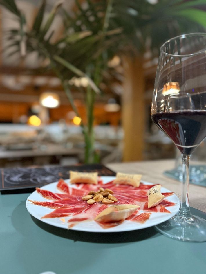 Plato de jamón ibérico con pan y frutos secos, copa de vino tinto en una mesa de restaurante.