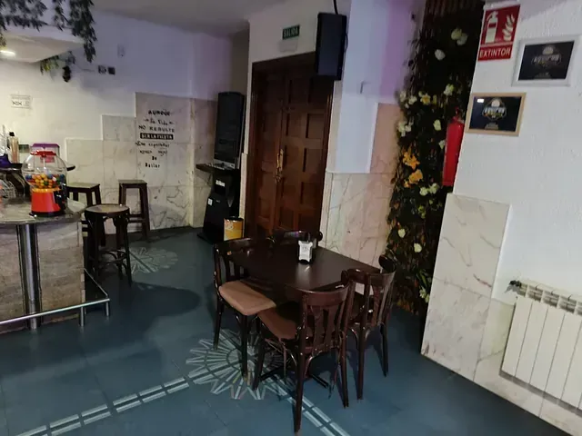 Interior de un bar con una mesa y sillas de madera oscura, una barra y una pared decorada con flores.