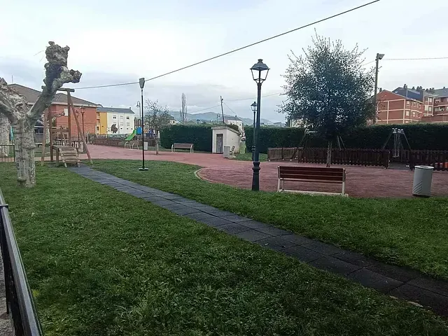Un parque con zona de juegos, bancos y un sendero, rodeado de césped verde y árboles. Al fondo se ven edificios y un cielo nublado.