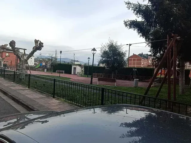 Parque con zona de juegos, bancos y árboles tras una valla negra y una calle. Cielo nublado.