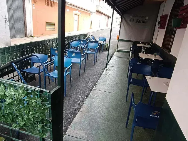 Zona de estar al aire libre de una cafetería con sillas y mesas azules. El espacio está parcialmente cubierto por un toldo, junto a una calle estrecha.