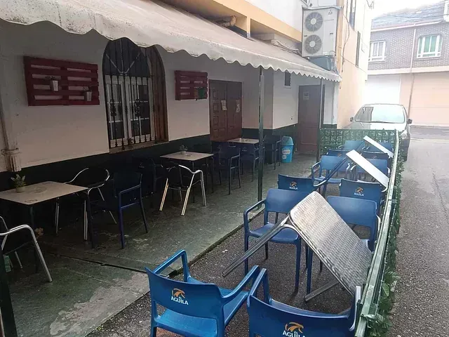 Exterior de un bar con sillas y mesas azules. Una mesa está volcada en una calle estrecha con un coche aparcado cerca.