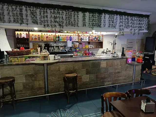 Interior de bar con barra de azulejos, taburetes y estanterías repletas de bebidas. Encima, enredaderas colgantes.