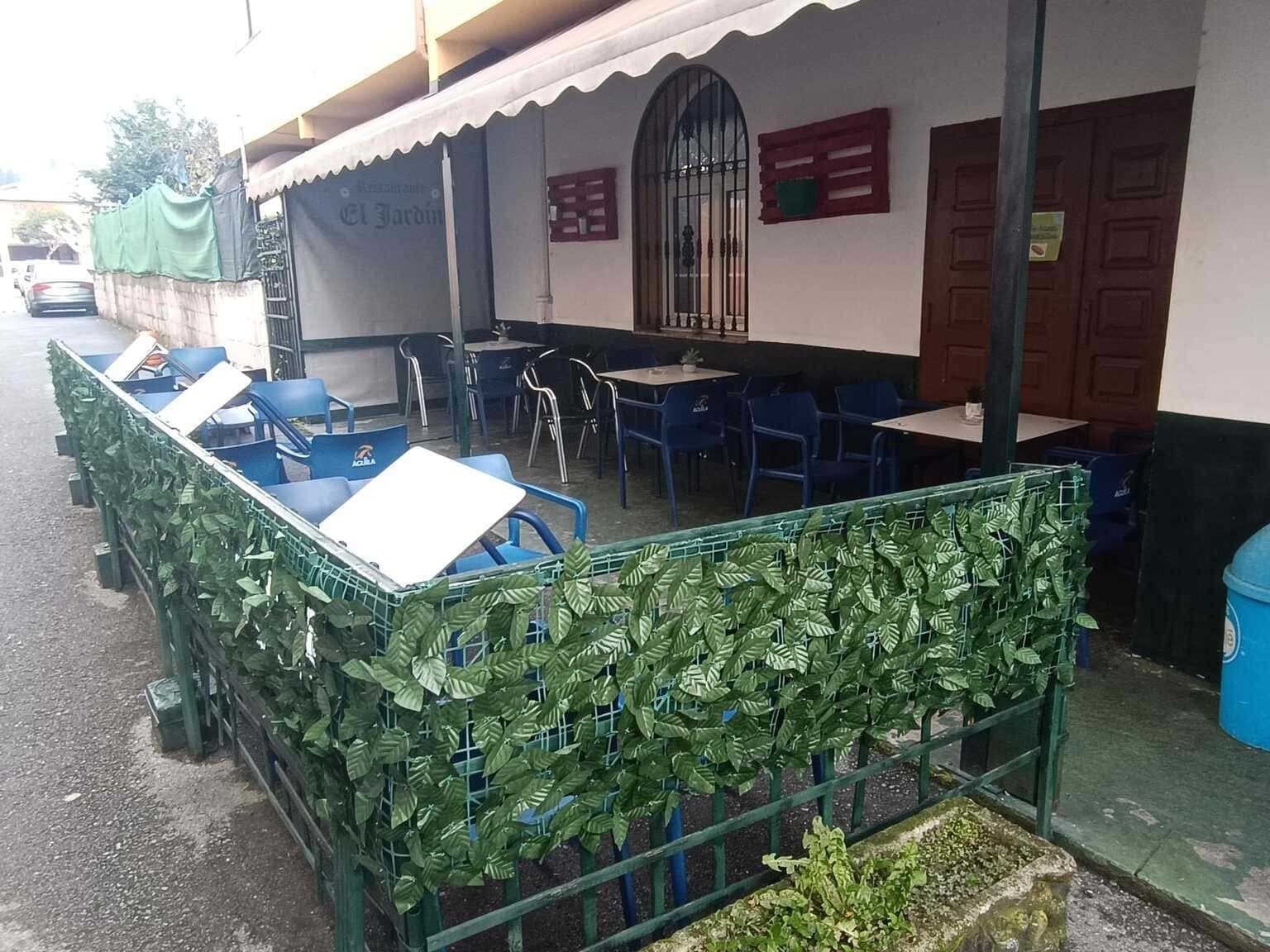 Una cafetería al aire libre con sillas y mesas azules, protegida por una valla verde con hojas artificiales. Un toldo blanco proporciona sombra.