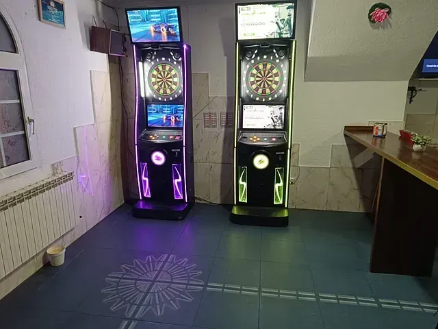 Dos máquinas de dardos iluminadas se encuentran en una habitación. El suelo tiene un diseño de rayos de sol y se ve una barra.