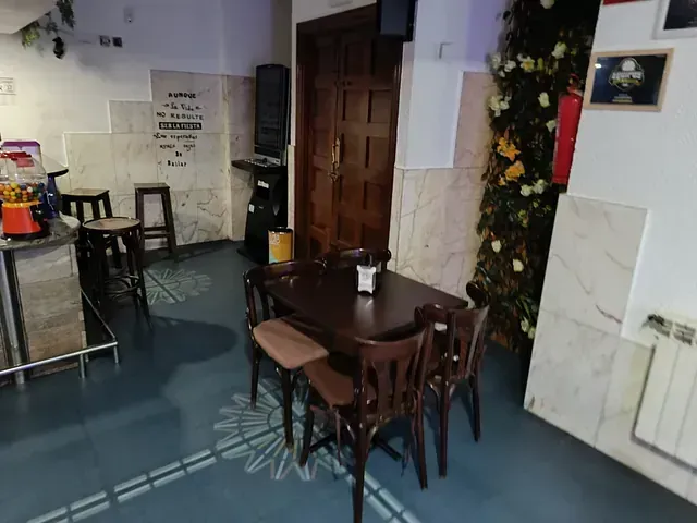 Interior de un bar con una mesa y sillas de madera oscura, un mostrador y una pared cubierta de flores.
