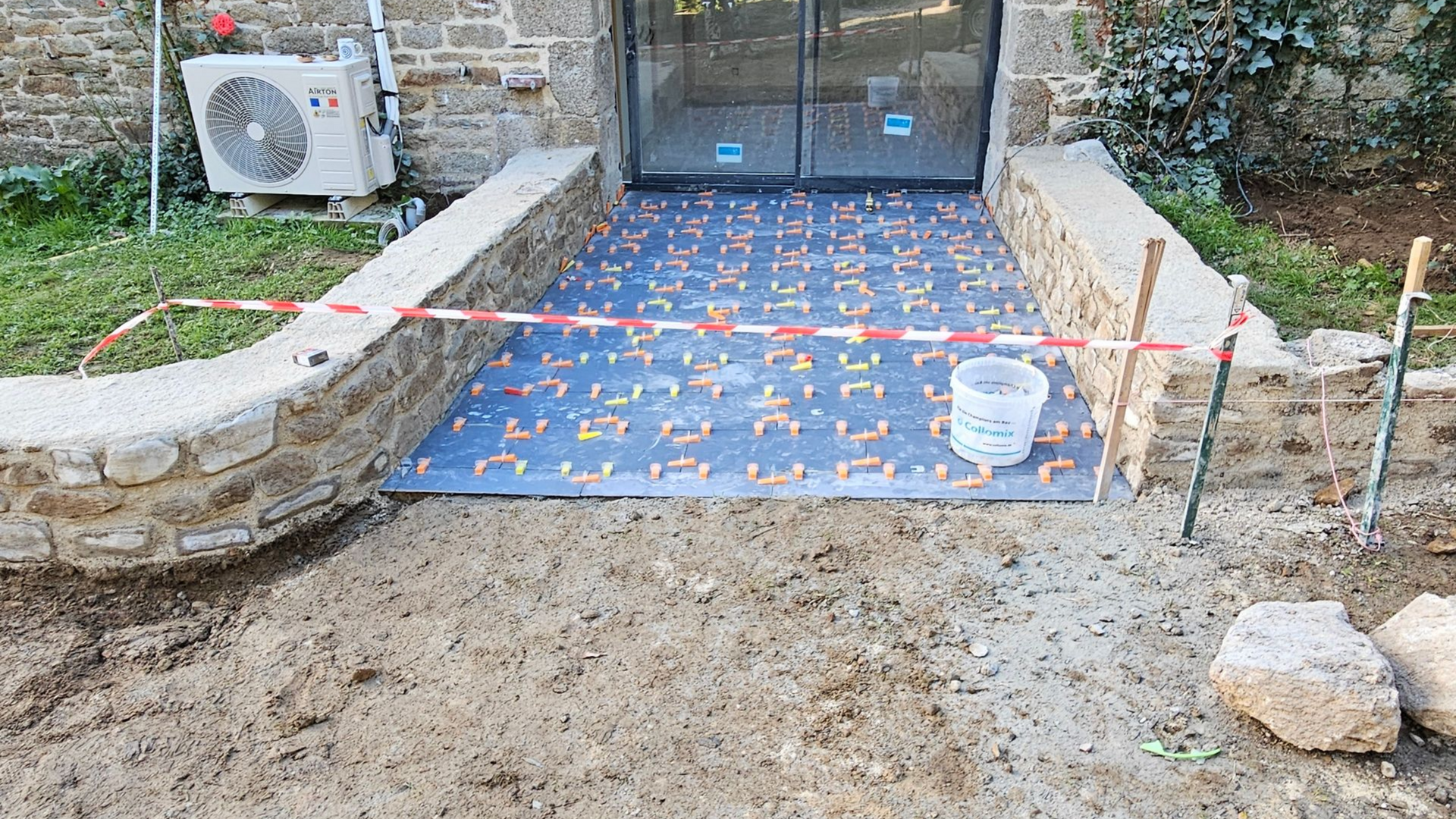 Construction d'une entrée avec pose de carrelage neuf, entourée de murs en pierre, de ruban de sécurité et d'outils.