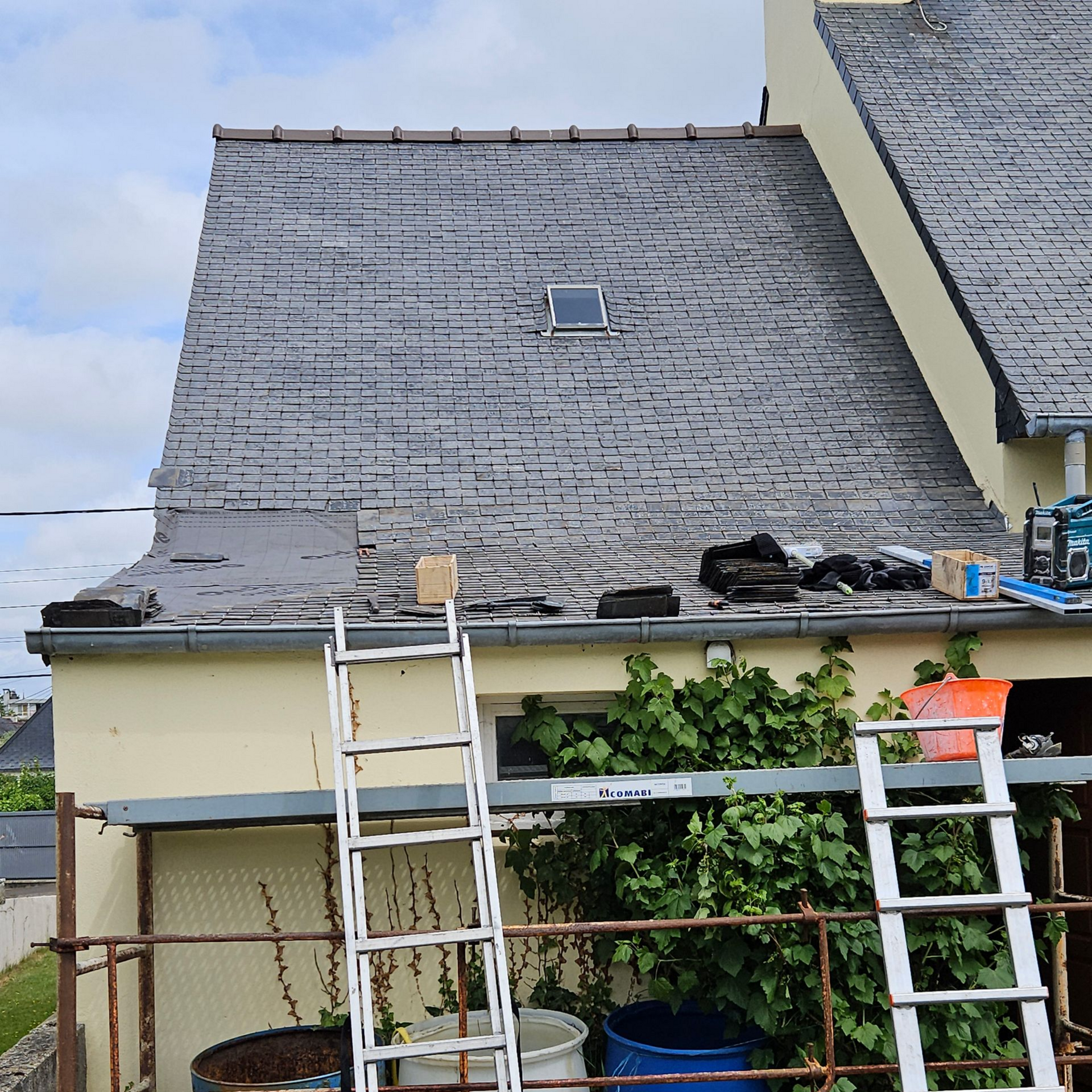 Travaux de réparation de toiture en cours : toit en tuiles grises, échelle, outils, puits de lumière, bâtiment beige.