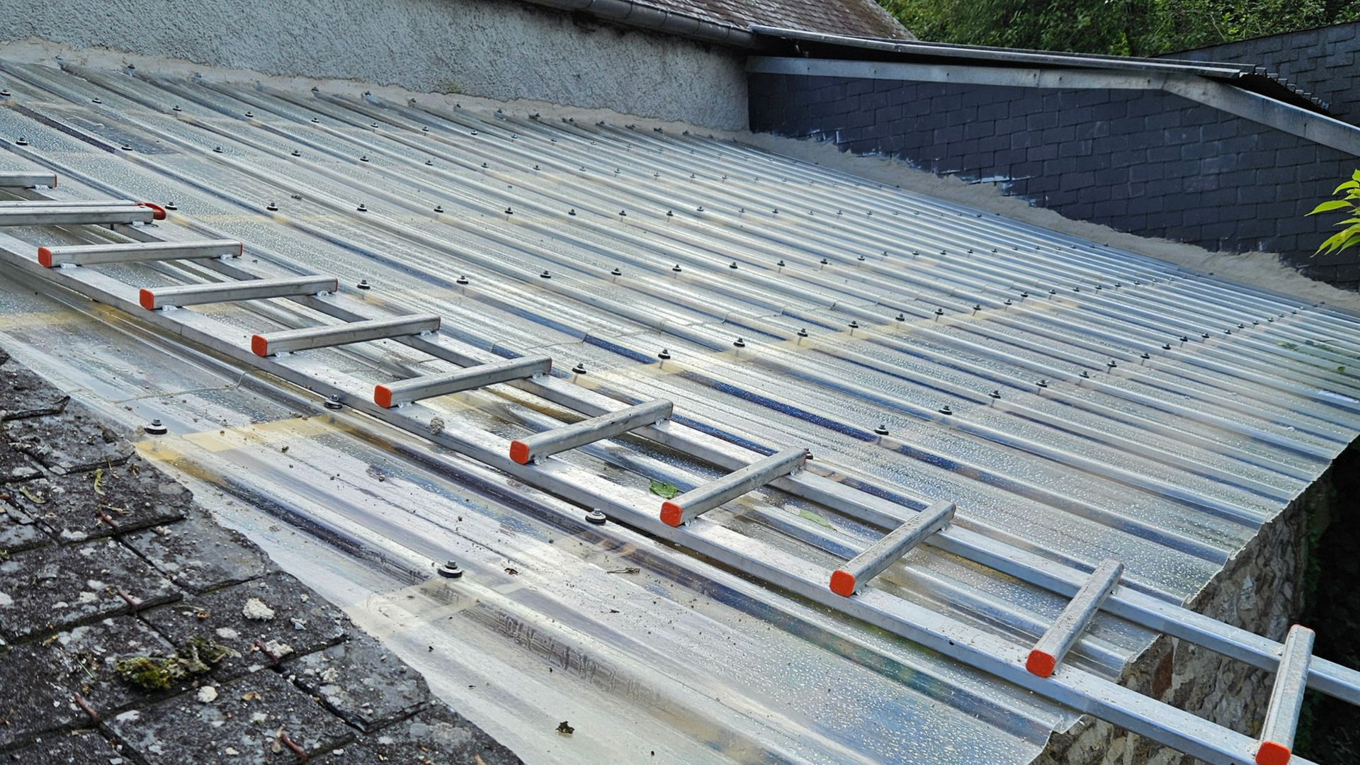 Échelle fixée sur un toit en tôle ondulée. Panneaux gris et transparents, laissant apparaître une partie du toit.