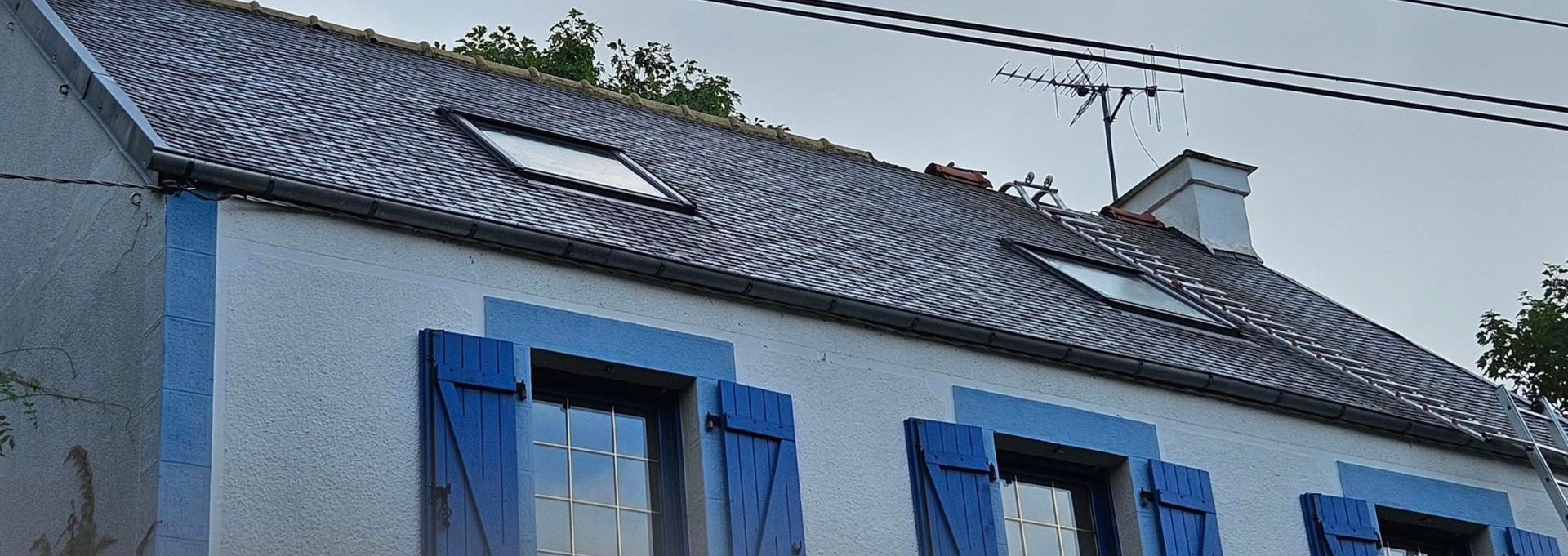 Une maison blanche avec des volets bleus, un toit sombre et deux puits de lumière.
