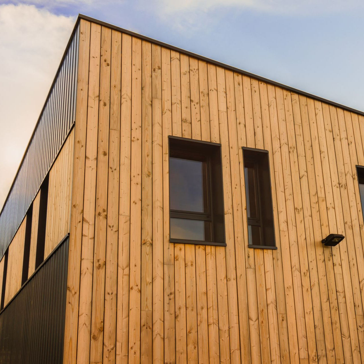 Extérieur d'un bâtiment en bois avec des panneaux de bois verticaux, des fenêtres à cadre sombre et une section partiellement visible à panneaux sombres.