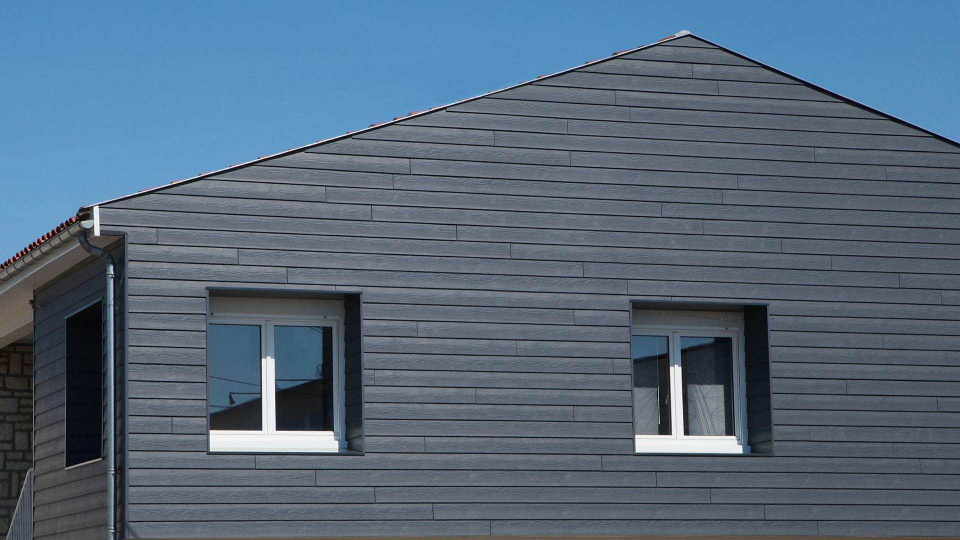 Bardage gris foncé sur une maison avec deux fenêtres à cadre blanc sous un ciel bleu.