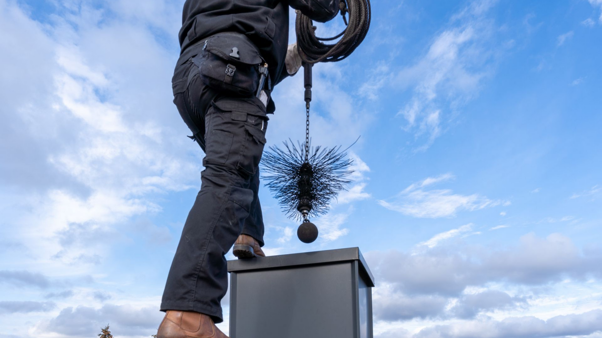 Ramoneur sur un toit, tenant brosse et câble contre un ciel nuageux.