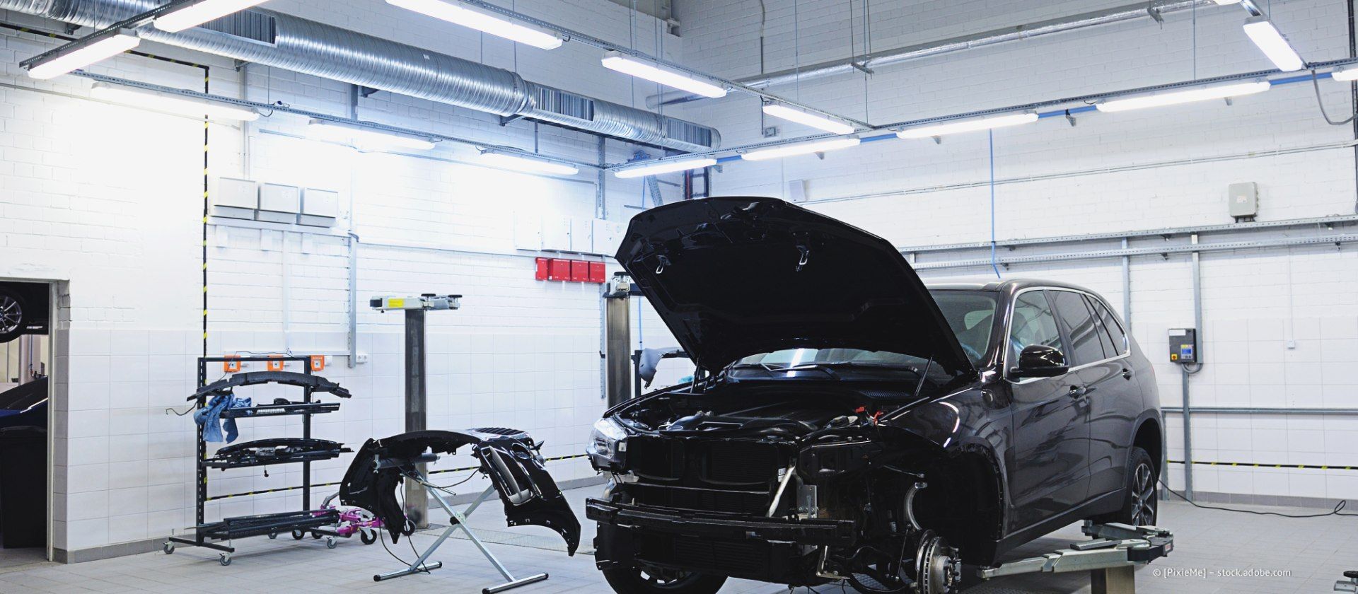Schwarzer SUV mit offener Motorhaube in einer hellen Autowerkstatt, die vordere Stoßstange ist abmontiert und Teile liegen verstreut daneben