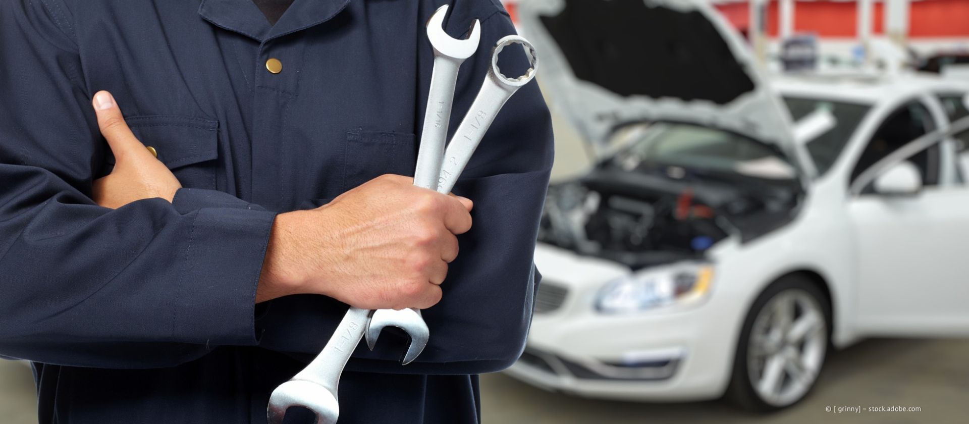 Ein Mechaniker hält Schraubenschlüssel und Schraubendreher in seiner Garage; im Hintergrund ist ein Auto zu sehen.